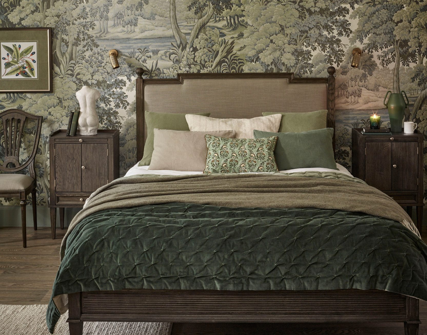 A cozy dramatic bedroom with a green and dark neutrals colour scheme featuring a dark wood bed with multiple pillows, Gustavian style side tables, and a nature-themed wallpaper.