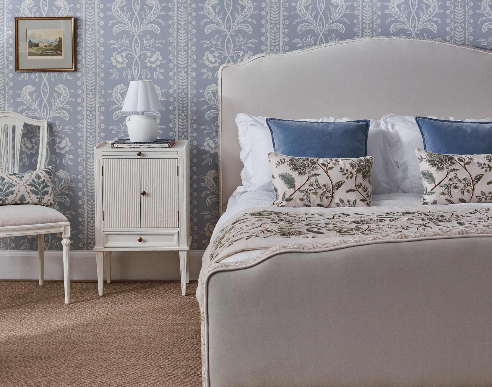 Elegant blue bedroom with a white upholstered bed, blue and floral cushions, a patterned throw, a white nightstand, and floral blue wallpaper.