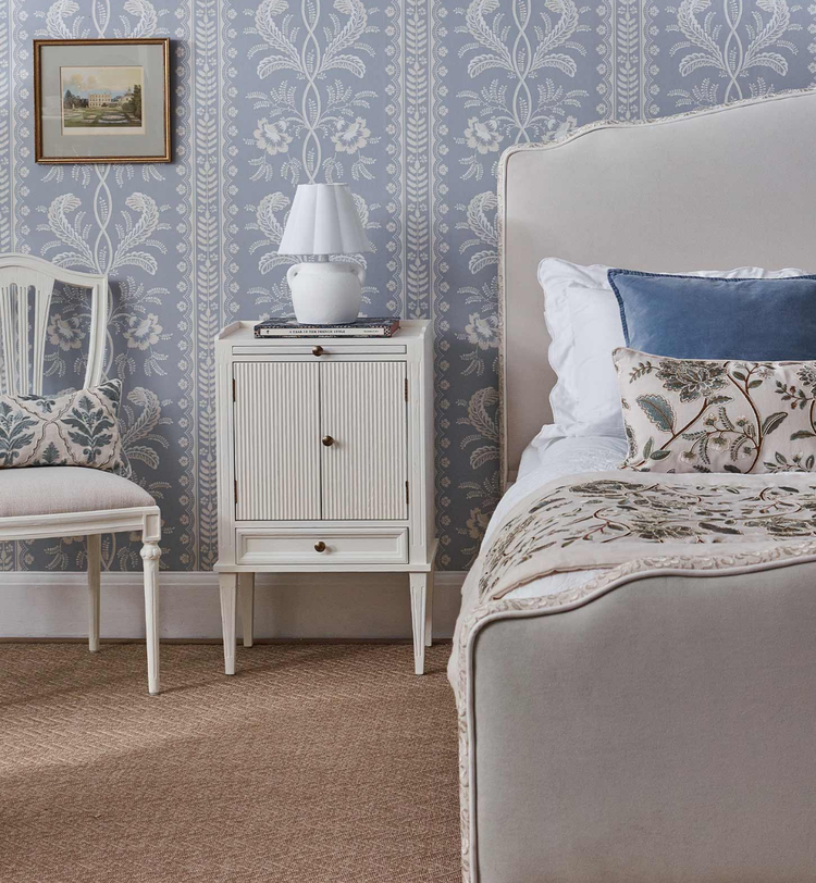 Elegant blue bedroom with a white upholstered bed, blue and floral cushions, a patterned throw, a white nightstand, and floral blue wallpaper.