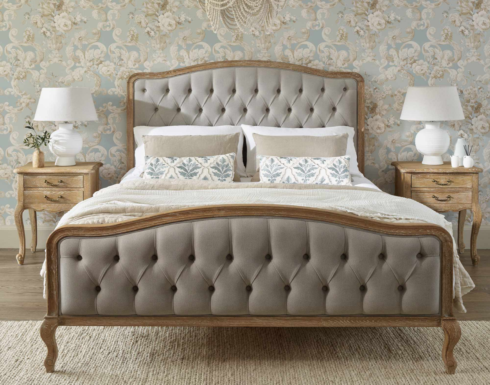 Elegant bedroom with a buttoned upholstered bed, floral wallpaper, two rustic wooden bedsides with white lamps, and a textured rug on the floor.