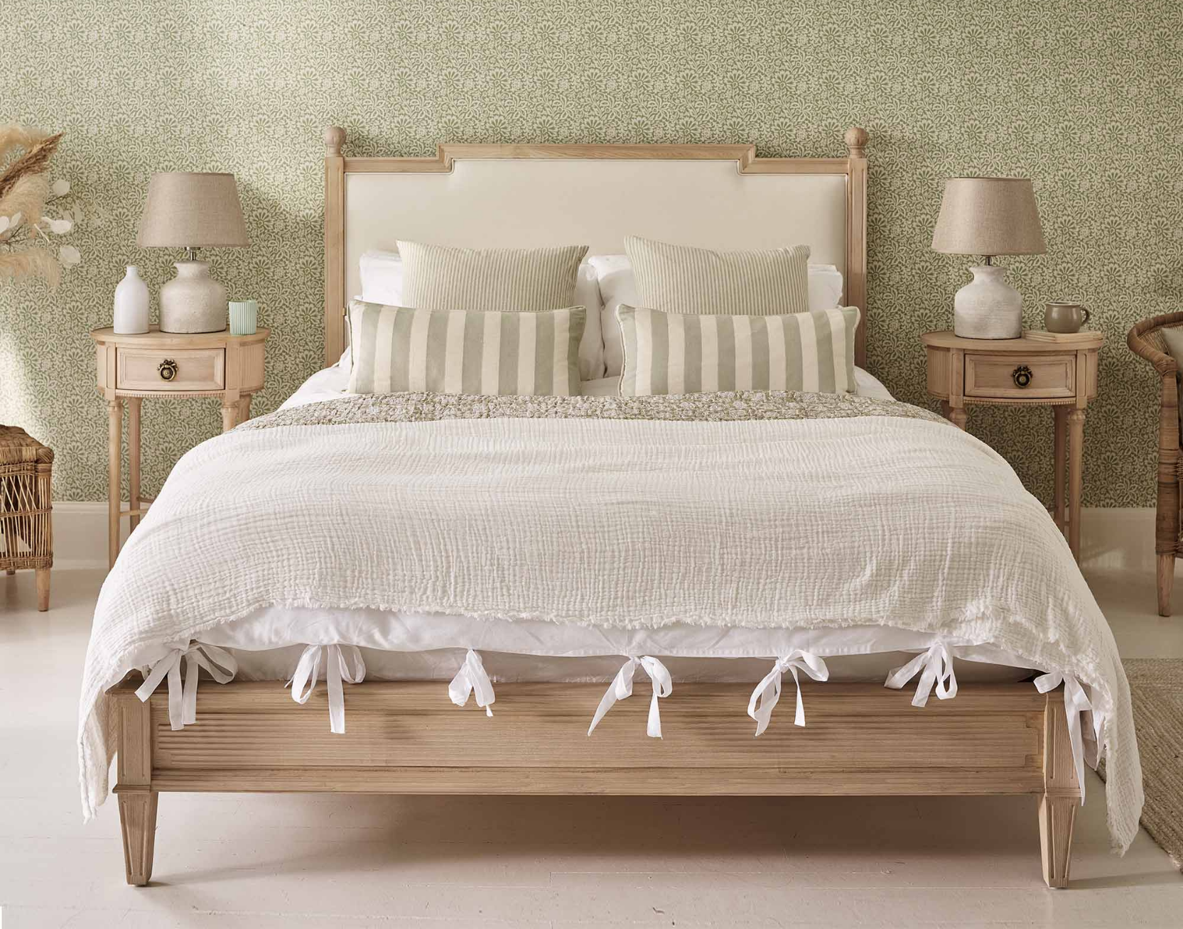 A cozy bedroom featuring a pale wooden bed with white bedding, striped cushions, and matching bedside tables with lamps, set against a patterned green wallpaper.