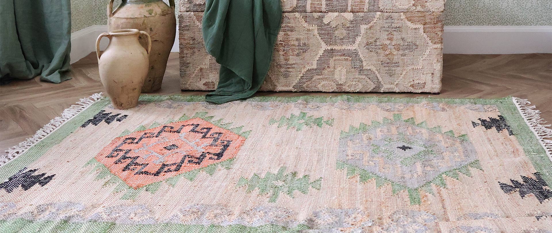 A woven rug with geometric patterns in orange, green, and gray on a wooden floor, near clay pots and a textured fabric-covered chest.