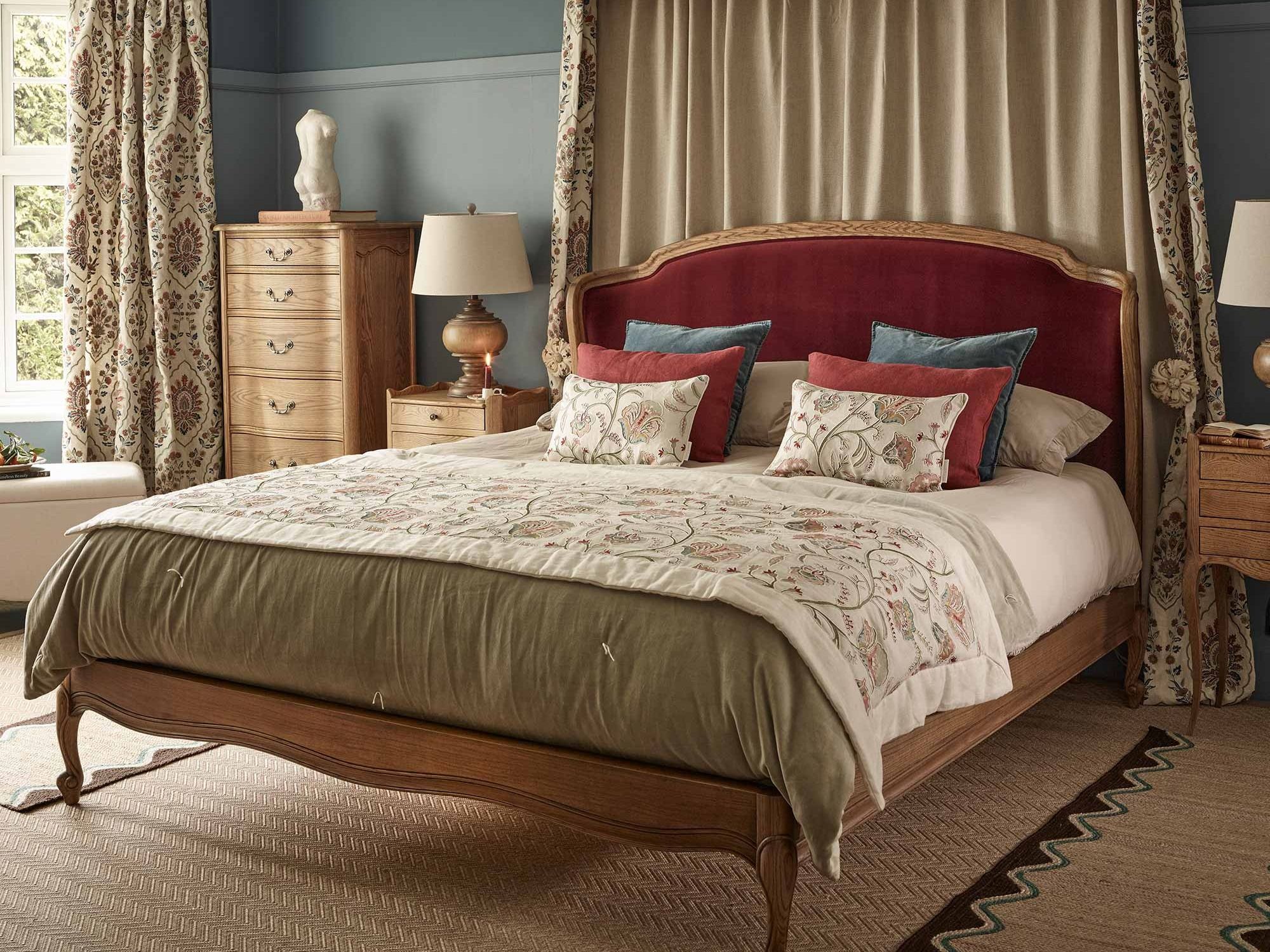 Elegant bedroom with a wooden bed, red velvet headboard, floral pillows and duvet. Decor includes vintage lamps, a sculpted bust, and patterned curtains.