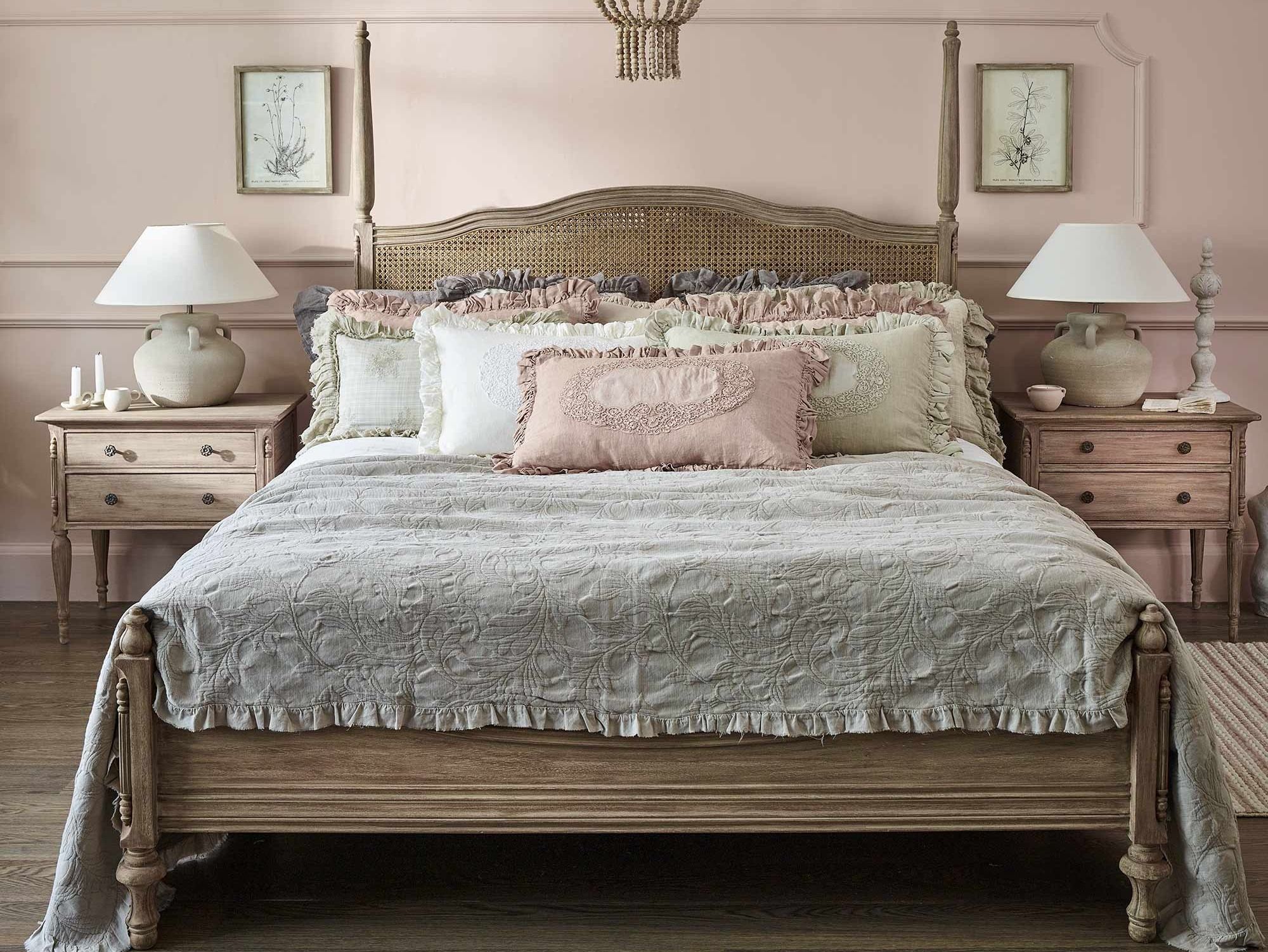 Vintage bedroom with a wooden bed, pastel ruffled bedding, matching nightstands, soft lighting, and framed botanical prints on pink walls.
