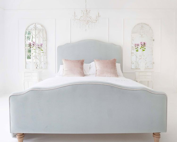 Elegant bedroom with a light gray upholstered bed, pink pillows, ornate mirrors, and a chandelier, creating a serene, airy atmosphere.