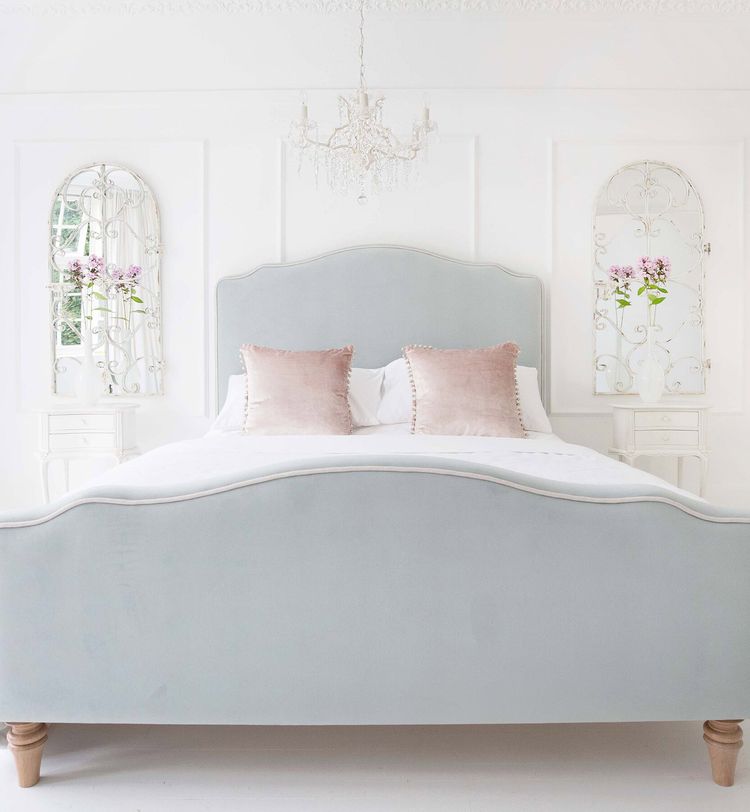 Elegant bedroom with a light gray upholstered bed, pink pillows, ornate mirrors, and a chandelier, creating a serene, airy atmosphere.