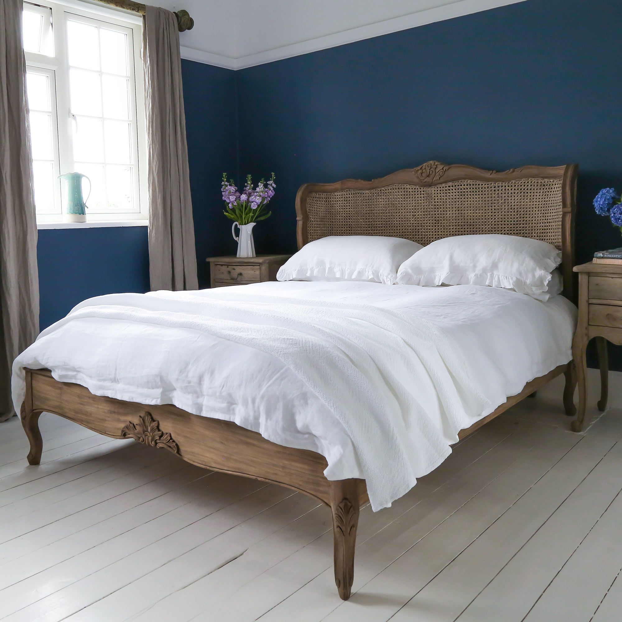 A cozy bedroom with a wooden bed, white bedding, blue walls, and natural light from a window. Two nightstands with flowers flank the bed.