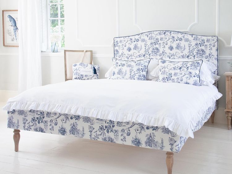 Elegant bedroom with a white and blue floral bedspread, matching pillows, and a large window. Light wood floors and a bird artwork on the wall.