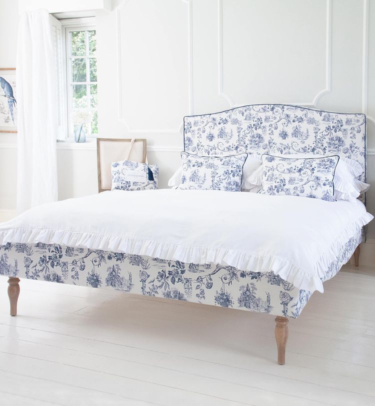 Elegant bedroom with a white and blue floral bedspread, matching pillows, and a large window. Light wood floors and a bird artwork on the wall.