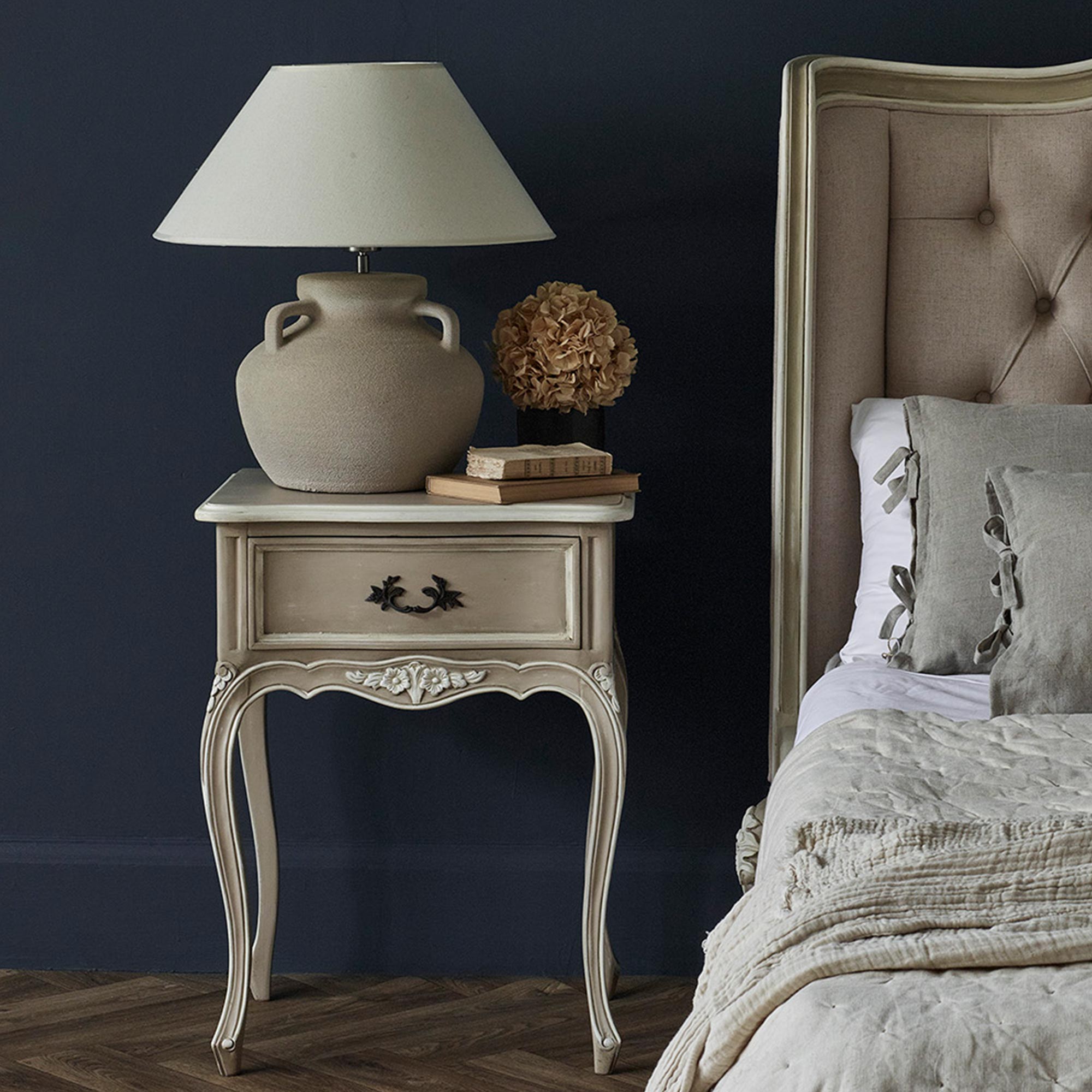 Elegant bedroom scene with a vintage nightstand, beige lamp, books, dried flowers, and a tufted bed against a dark wall.