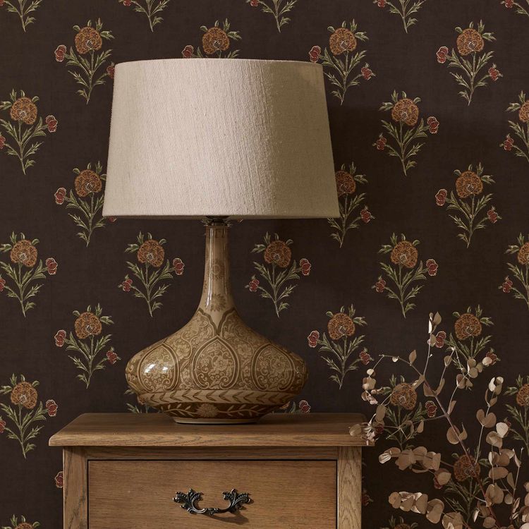 Ornate ceramic lamp with a beige shade on a wooden nightstand, set against floral-patterned wallpaper in earthy tones.