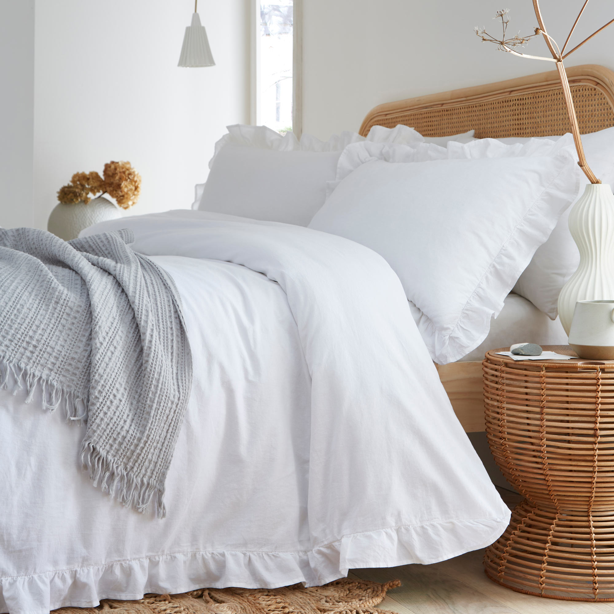 Cozy bedroom with a white ruffled bedding set, light gray throw, wicker side table, and decorative vase on a wooden floor.