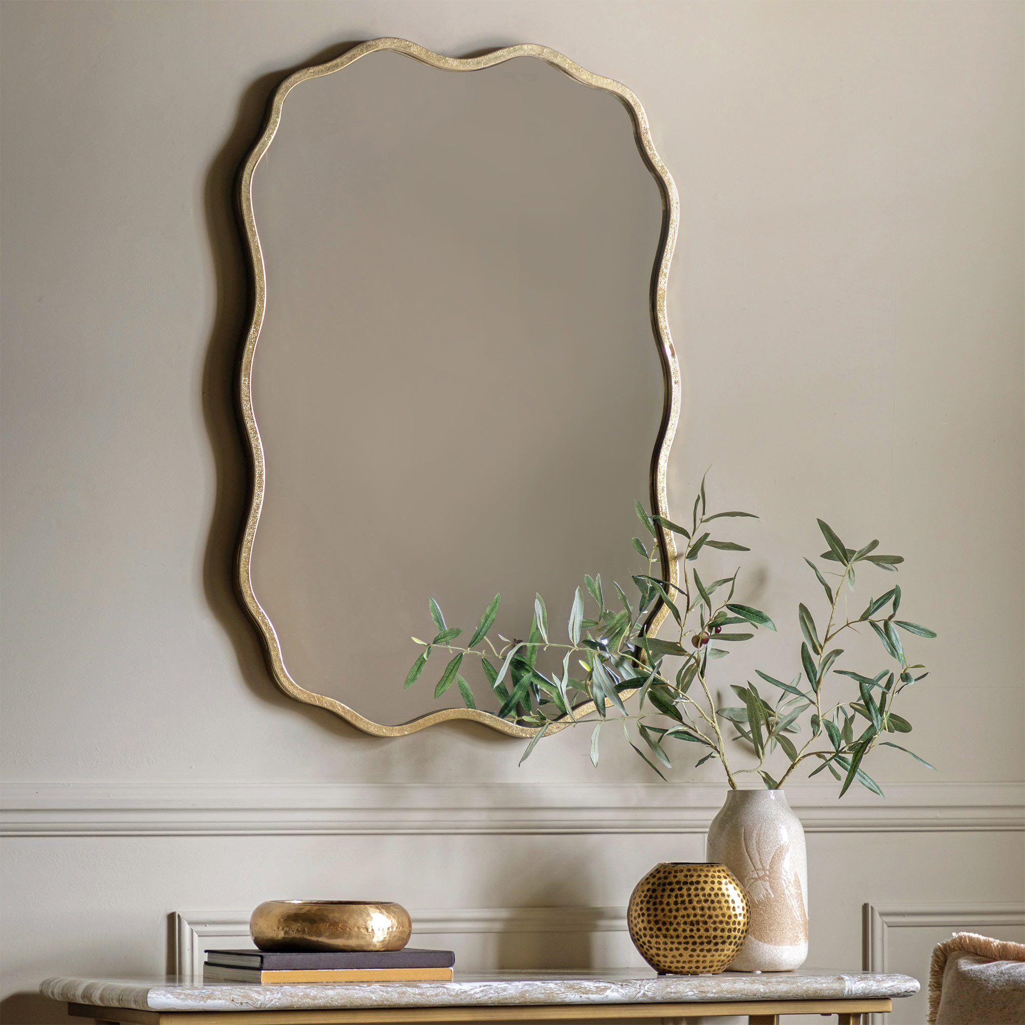 Elegant mirror with a wavy gold frame hangs above a marble table with decorative vases and green branches, set against a light-colored wall.