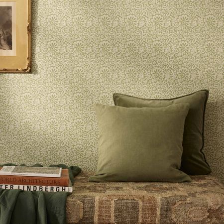 Two green velvet pillows on a patterned bench, with a textured green wallpaper and a framed picture partially visible on the wall.