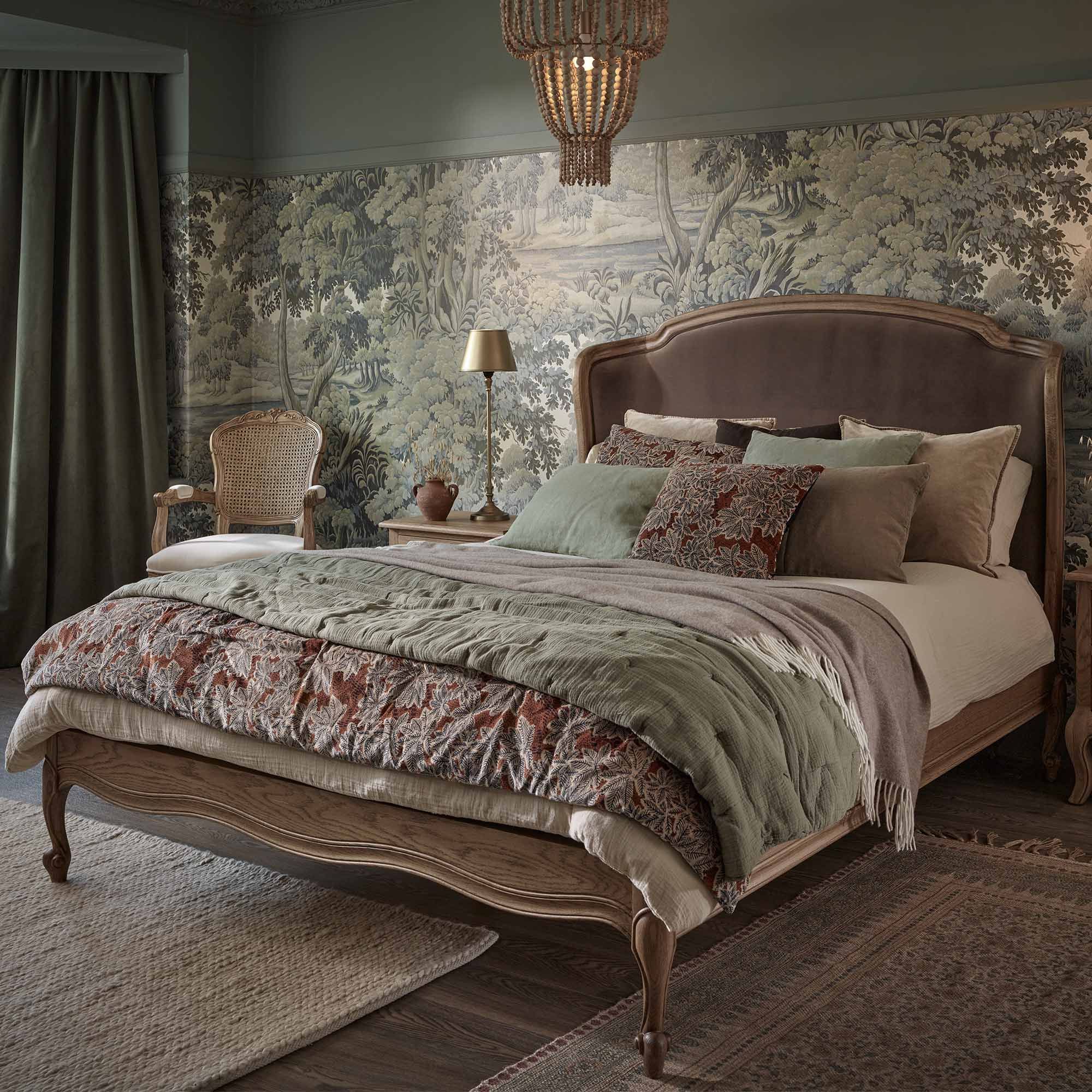 Elegant bedroom with a vintage bed, patterned bedding, and a tapestry-like wall. A woven chair and lamp sit by the bed. Warm, earthy tones prevail.