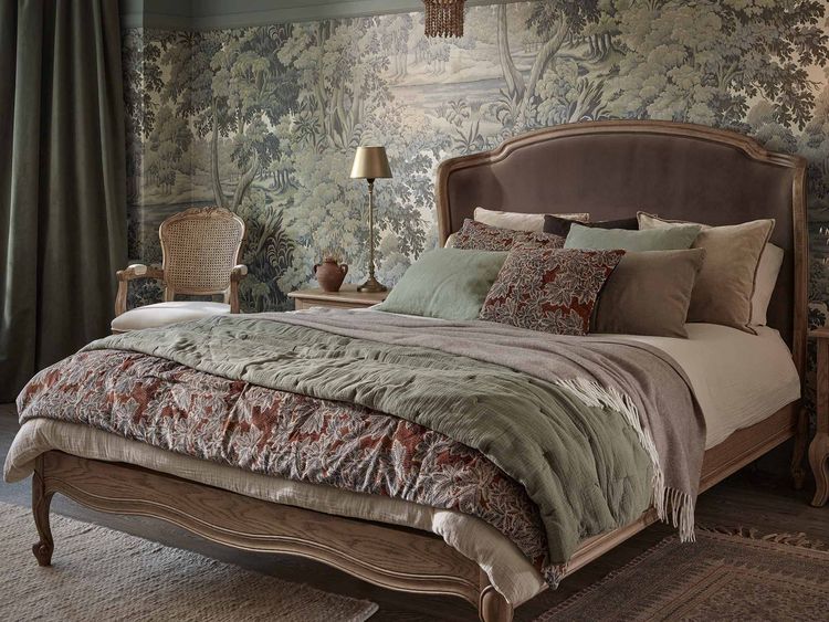 Elegant bedroom with a vintage bed, patterned bedding, and a tapestry-like wall. A woven chair and lamp sit by the bed. Warm, earthy tones prevail.