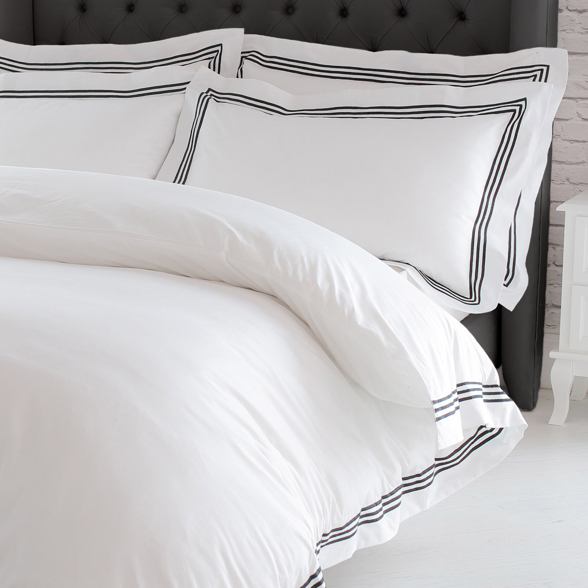 White bed with black-striped pillows and duvet, against a tufted gray headboard and a white bedside table.