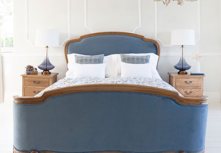 Elegant bedroom with a blue velvet bed, ornate wooden frame, two bedside tables with lamps, and a chandelier against a white panelled wall.