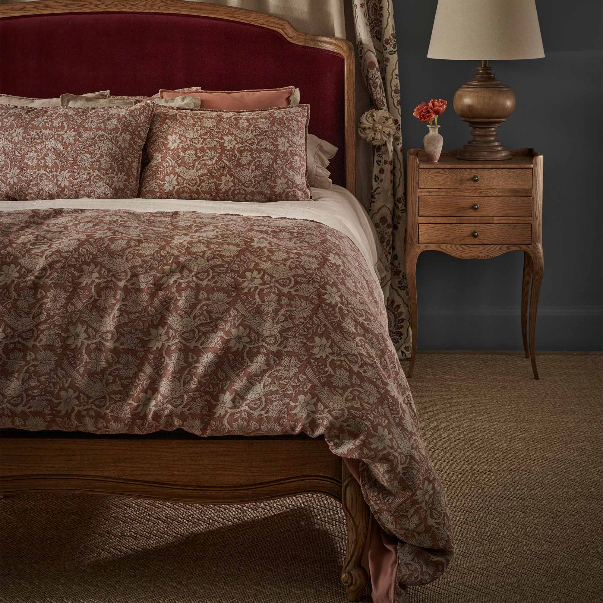 Elegant bedroom with a patterned bedspread in terracotta, velvet upholstered headboard, and a vintage nightstand holding a lamp and vase with flowers.