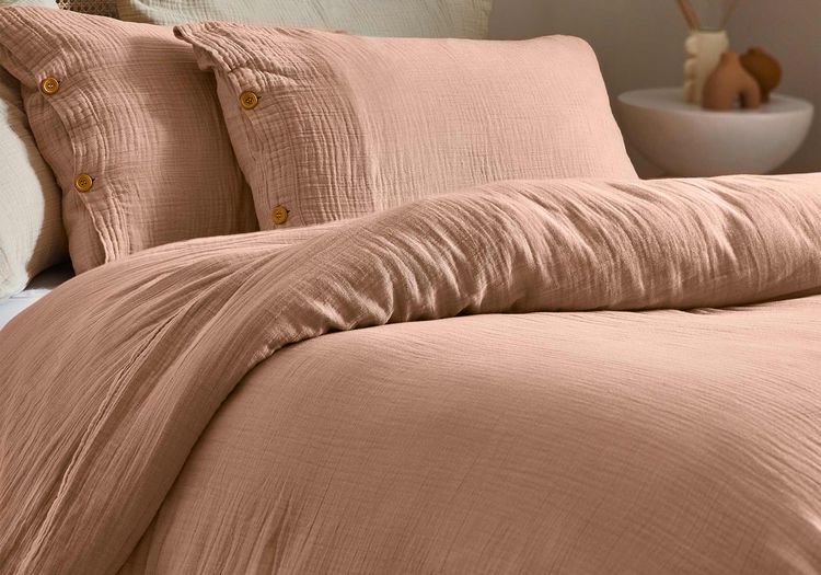 Cozy bed with soft, textured peach bedding and buttoned pillows, set against a light wood headboard. Neutral decor in the background.