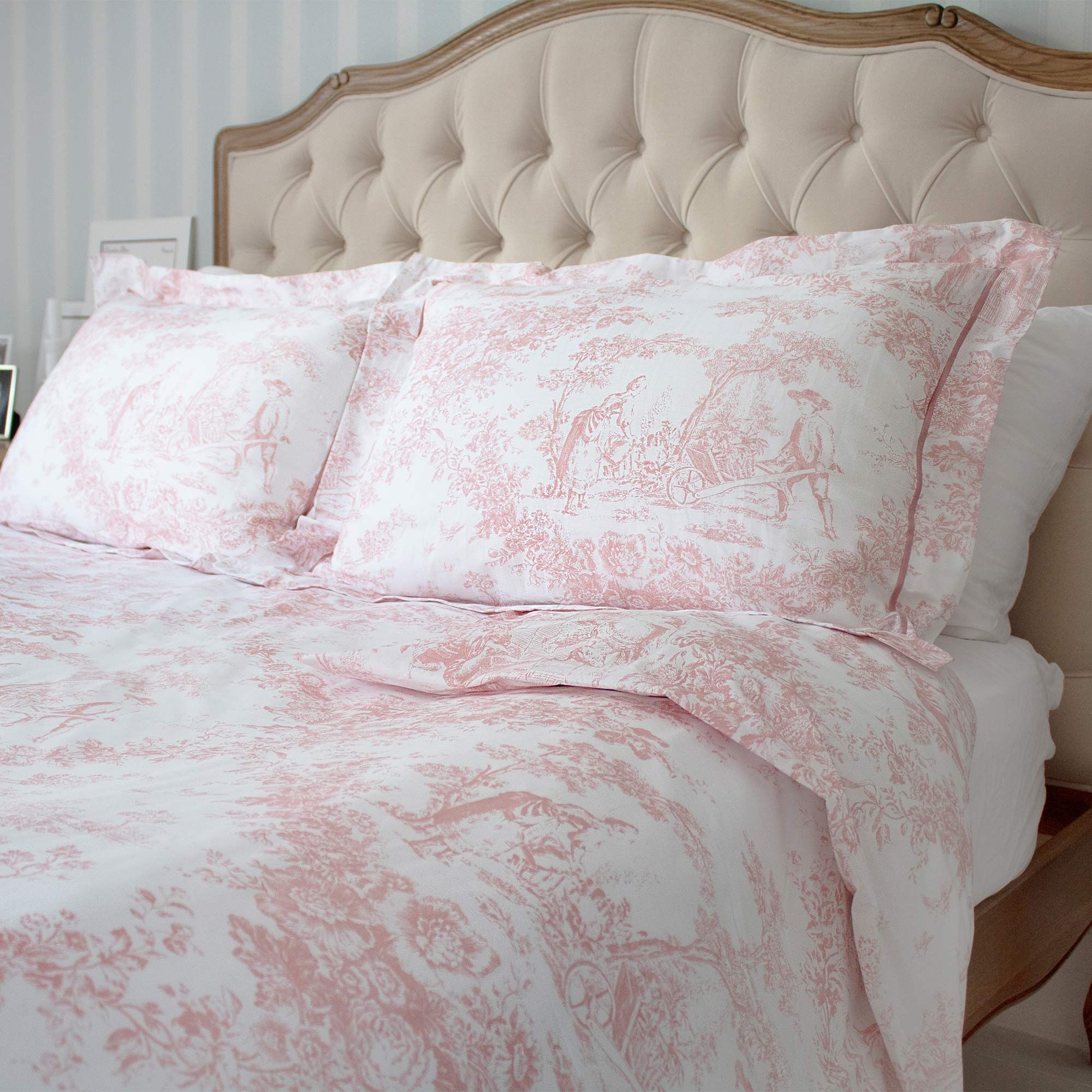Elegant bed with a tufted headboard, adorned with pink and white patterned bedding featuring pastoral scenes, in a softly lit bedroom.