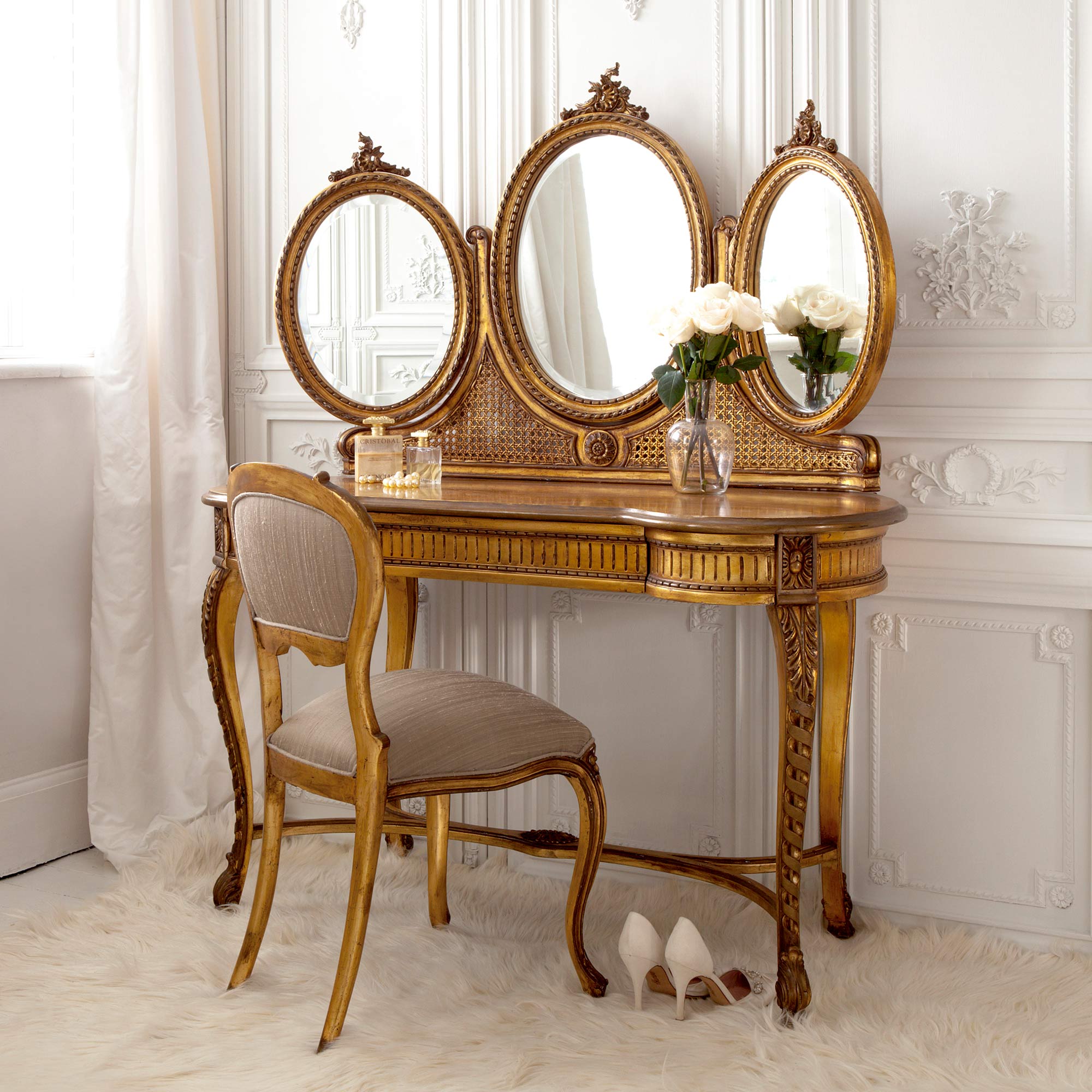 Elegant vanity with triple oval mirrors, ornate gold detailing, cushioned chair, roses in a vase, perfume bottles, and white heels on a plush rug.