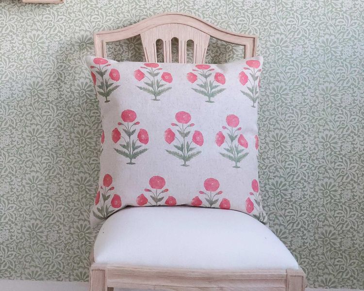A wooden chair with a white seat holds a cushion featuring pink floral patterns, set against a green floral wallpaper background.