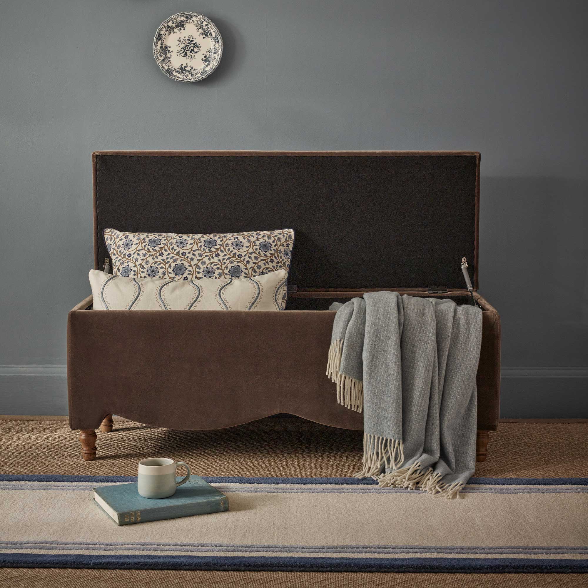 Open storage ottoman bench with patterned cushions and a sky blue throw, beneath a decorative plate on a blue wall.