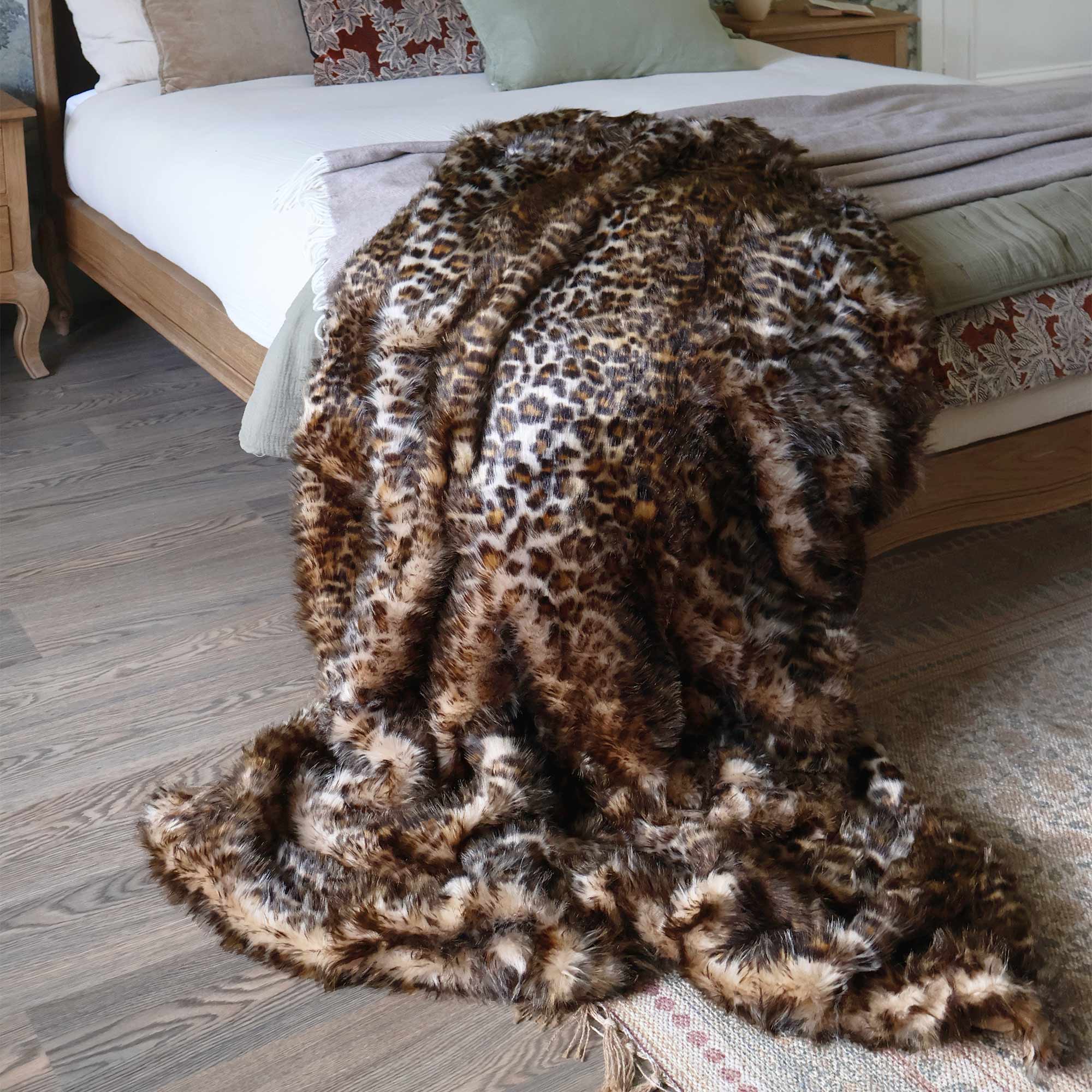 A leopard-print fur throw draped over the edge of a bed in a cozy bedroom with wooden flooring and neutral-toned decor.
