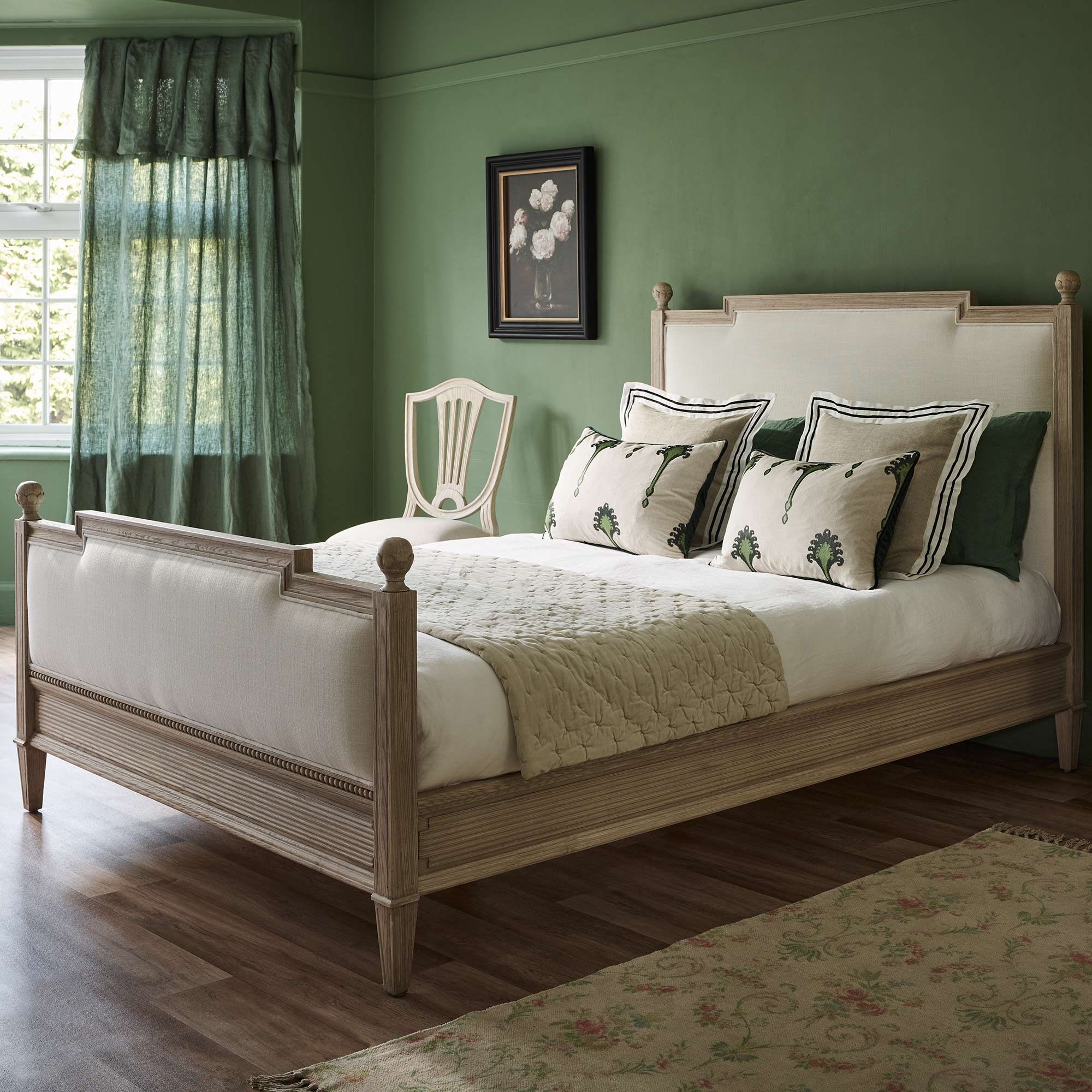 Elegant bedroom with a wooden bed, green walls, floral pillows, and a quilted blanket. A chair and framed artwork add charm. Natural light filters in.