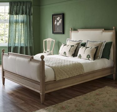 Elegant bedroom with a wooden bed, green walls, floral pillows, and a quilted blanket. A chair and framed artwork add charm. Natural light filters in.