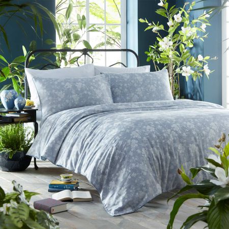 A cozy bed with floral-patterned blue bedding in a bright room, surrounded by lush green plants and scattered books on the floor.