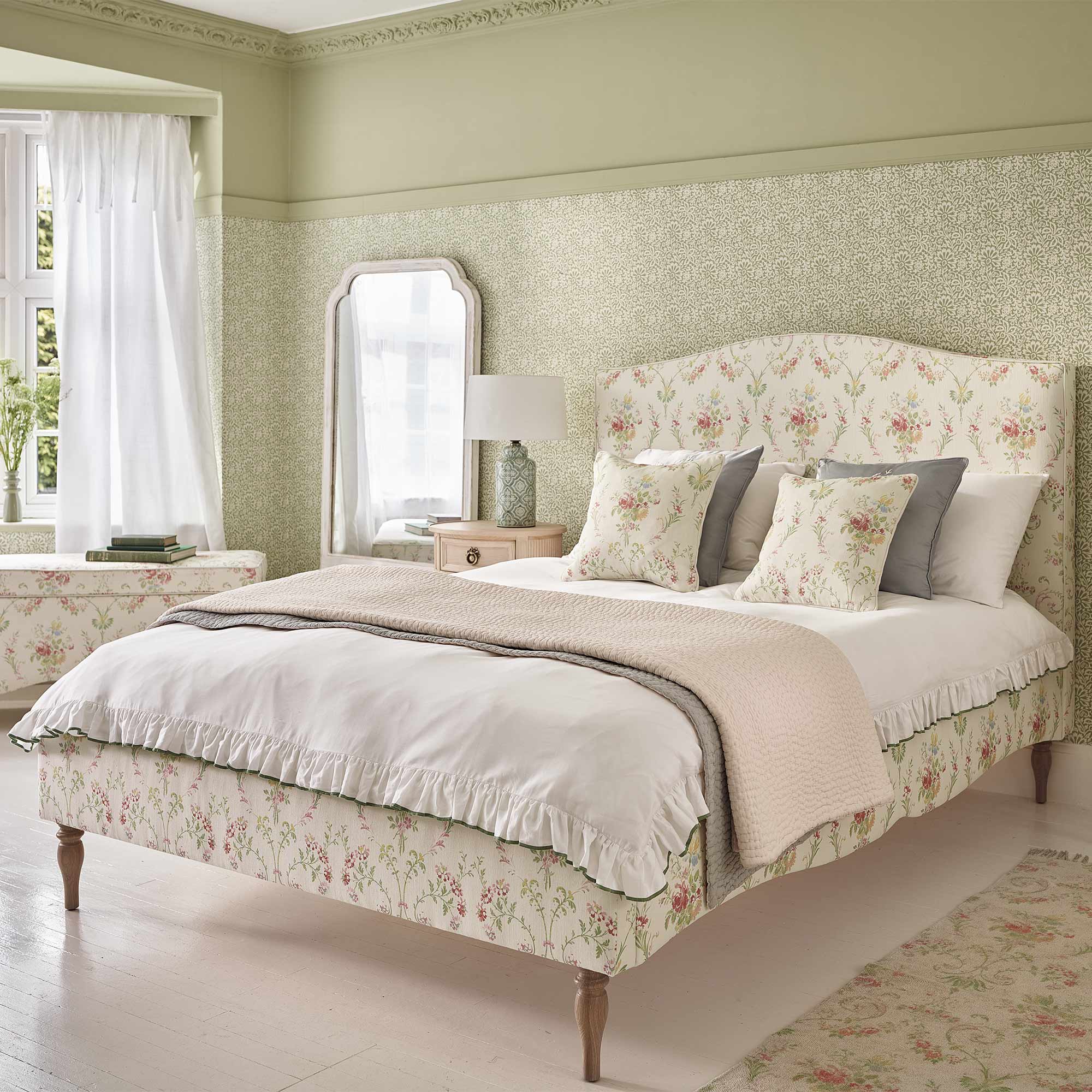 A cozy bedroom with a floral-patterned bed, decorative pillows, a bedside table with a lamp, a mirror, and soft green walls.
