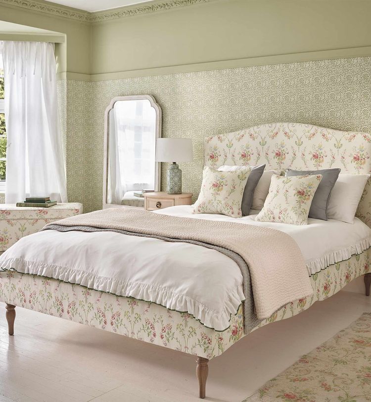 A cozy bedroom with a floral-patterned bed, decorative pillows, a bedside table with a lamp, a mirror, and soft green walls.