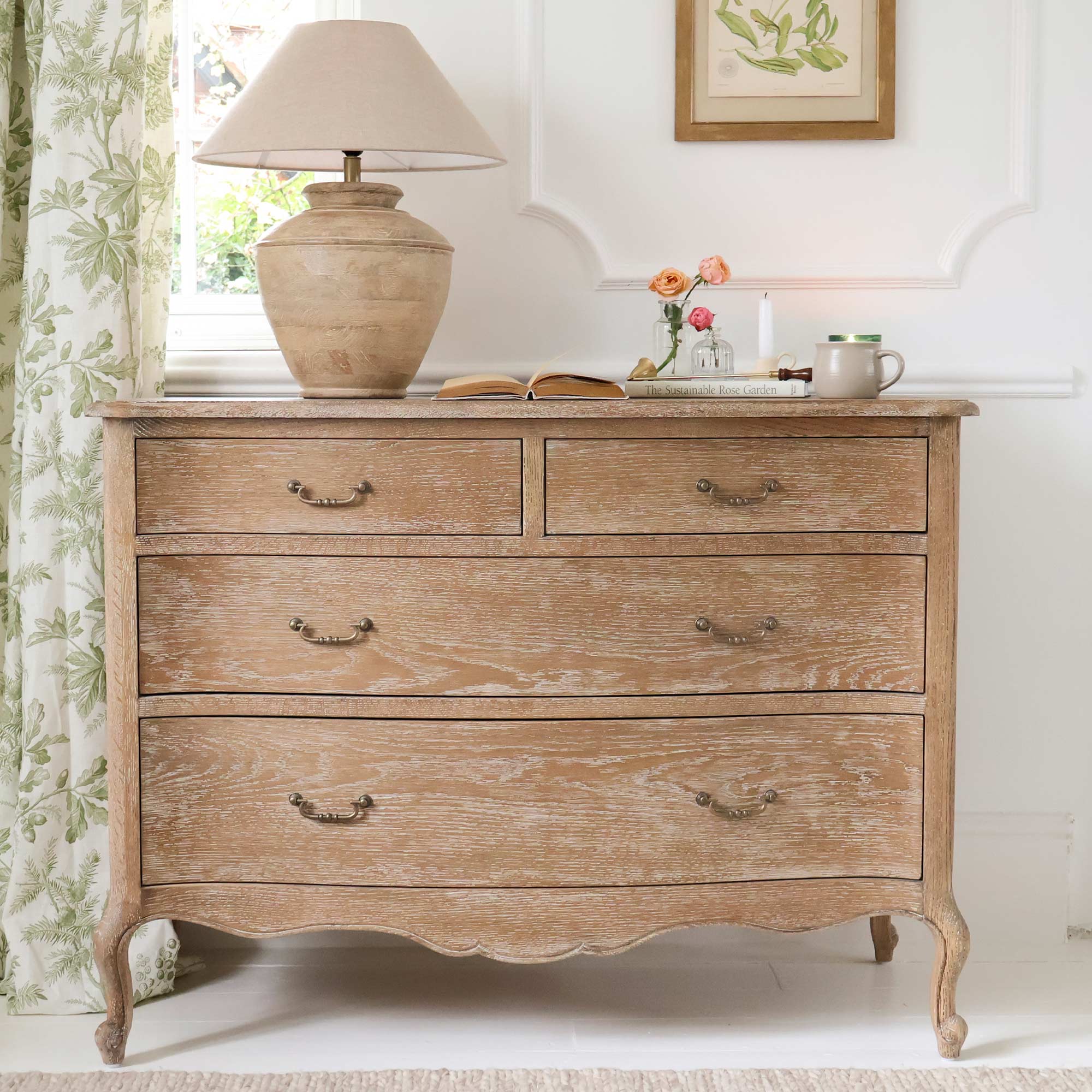 French-style wooden dresser with four drawers, topped with a lamp, open book, flowers, and a mug. Framed art hangs above; floral curtain on the side.