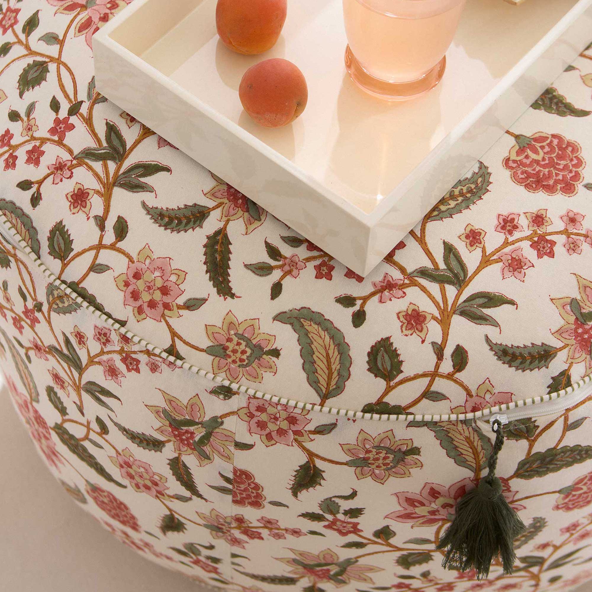 Floral-patterned ottoman with a white tray holding apricots and a glass of pink drink, featuring a decorative tassel.