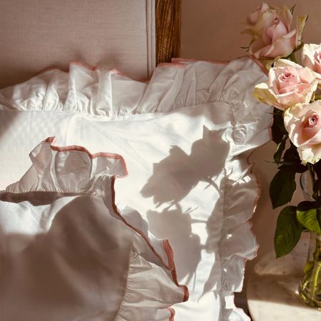 White ruffled pillows with pink trim on a chair, next to a vase of pink roses casting a shadow, in soft natural light.
