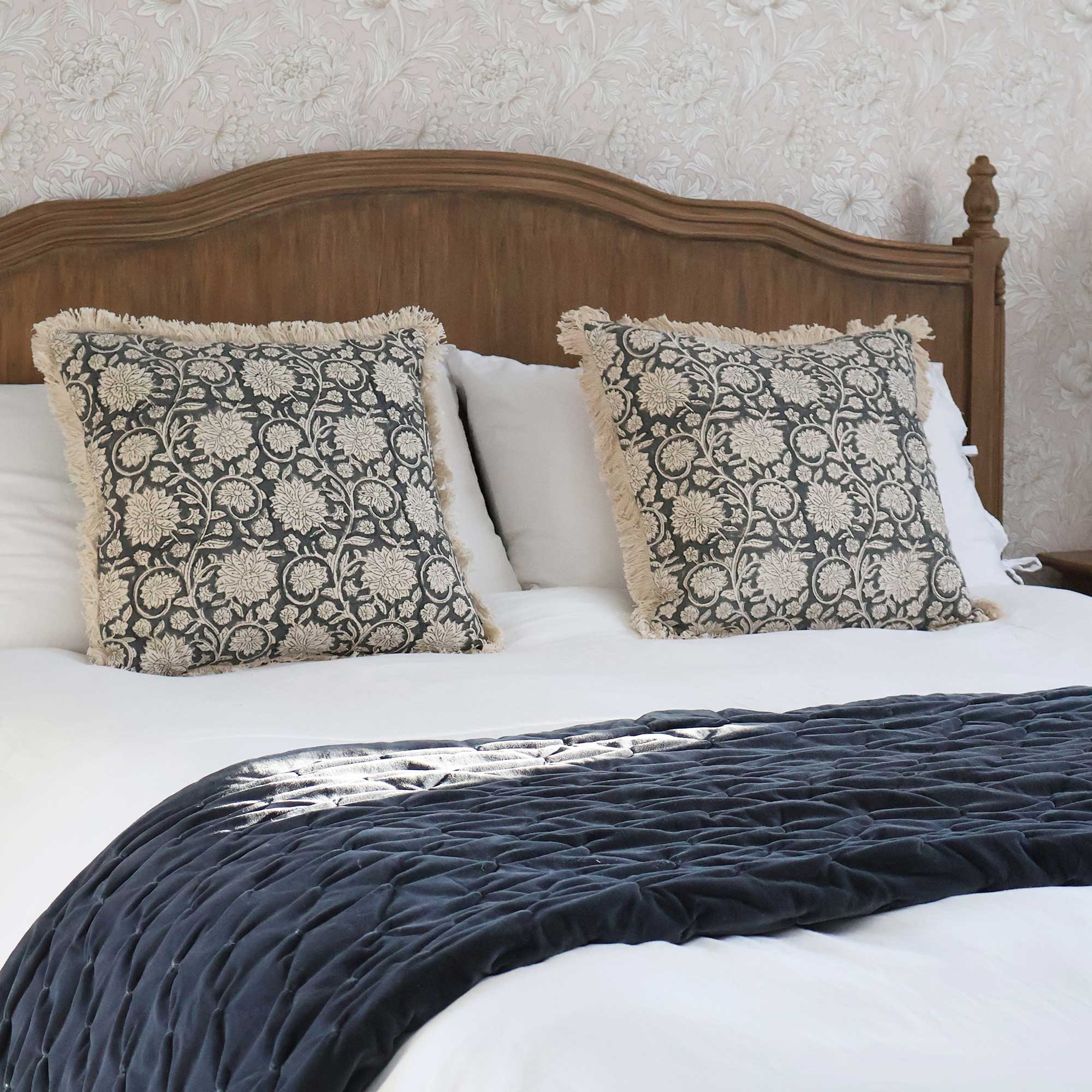 A neatly made bed with patterned pillows, a dark quilted throw, and a wooden headboard against a floral wallpaper background.