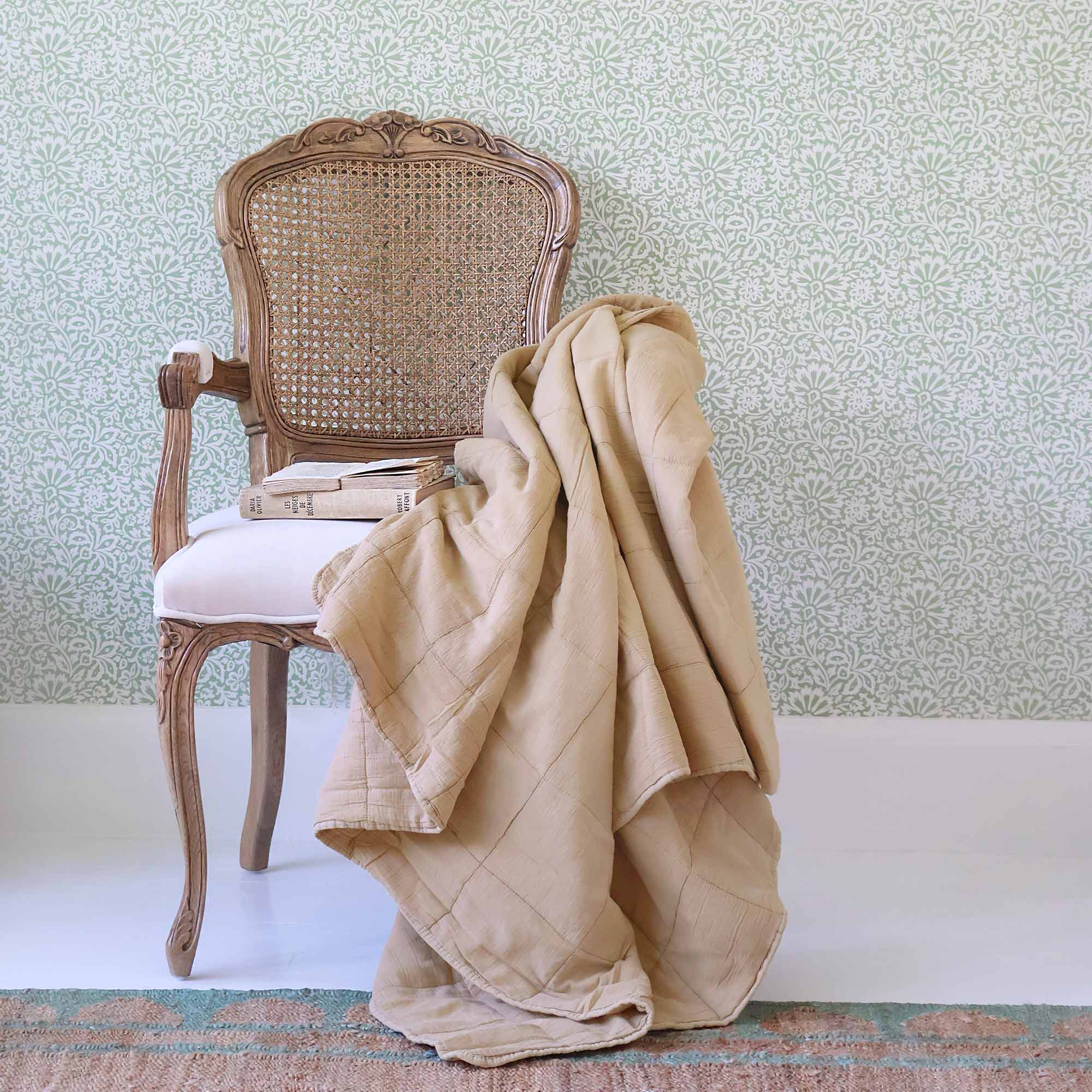 Ornate wooden chair with a rattan wicker backrest, draped with a beige quilt, on a jute rug.