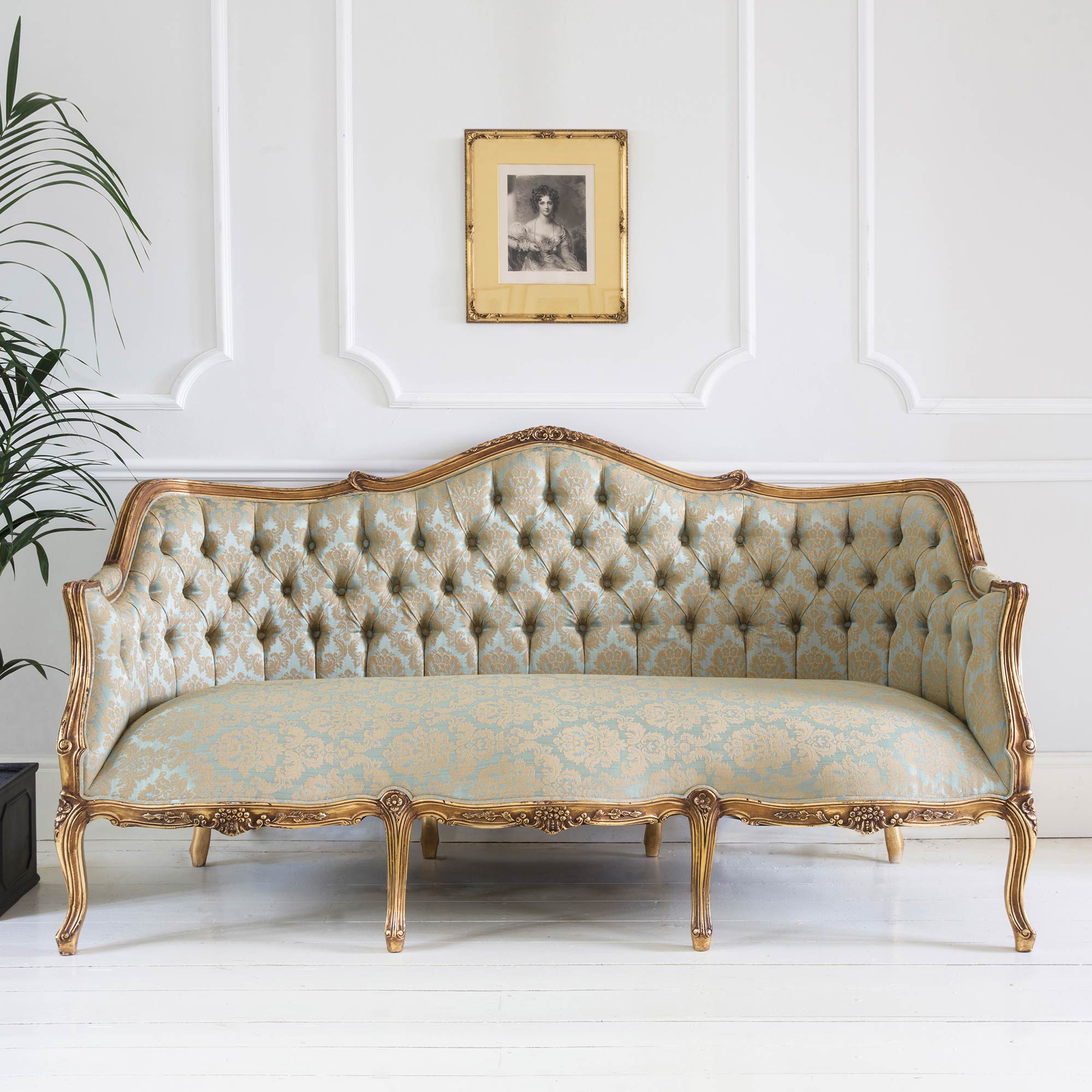 Elegant vintage sofa with ornate gold frame and tufted upholstery in a light blue pattern, set against a white wall with a framed picture.