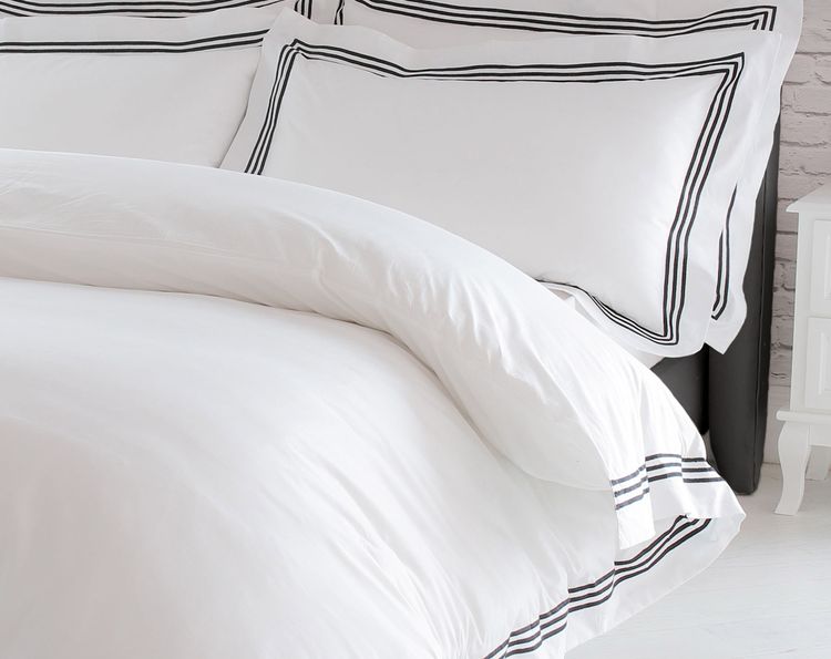 A neatly made bed with white bedding featuring black striped borders, set against a dark tufted headboard and a white bedside table.