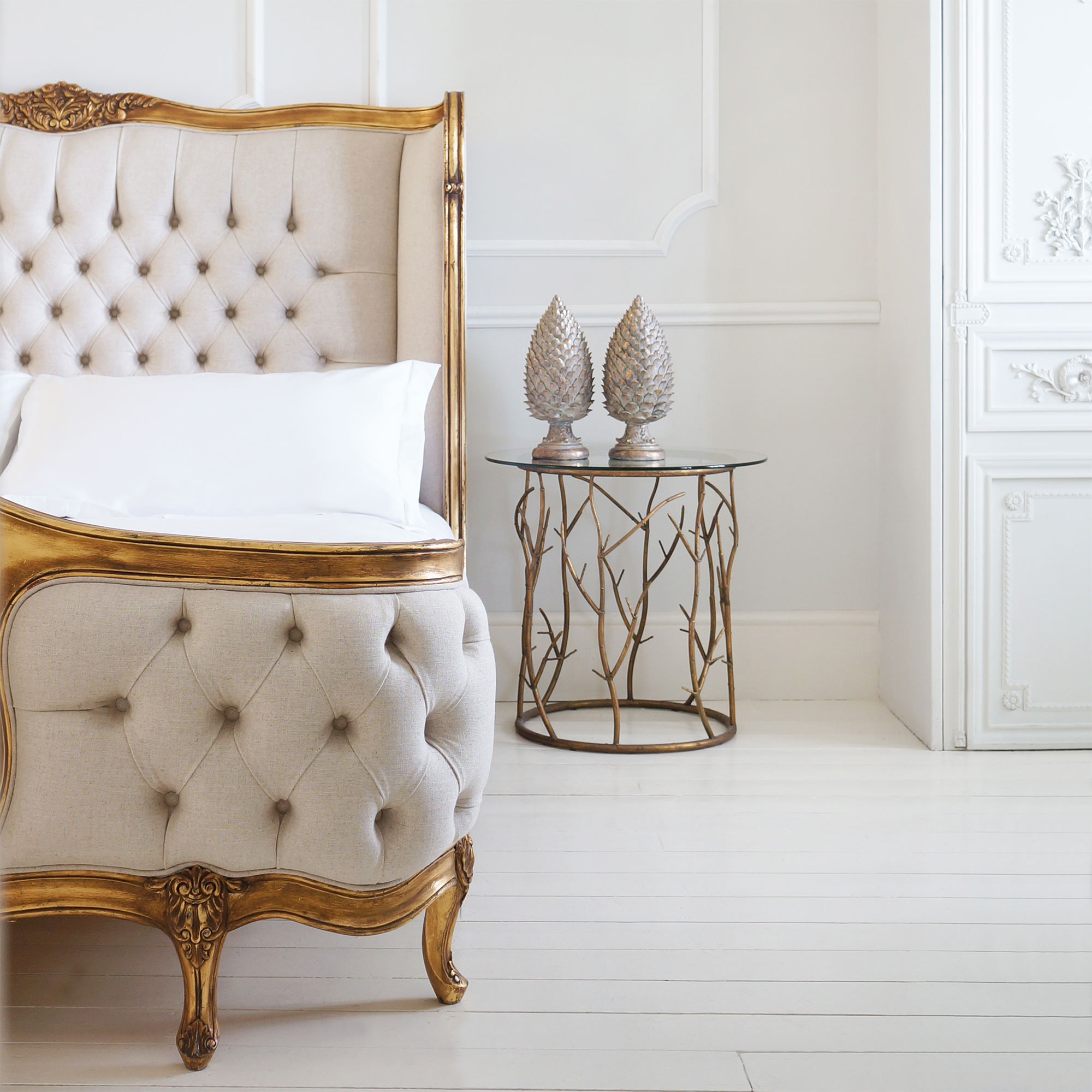 Elegant bedroom with a tufted beige bed, ornate gold frame, and a glass side table with decorative pineapples on a white wooden floor.