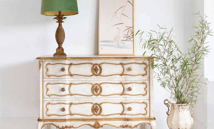 Vintage dresser with ornate handles, topped with a pineapple lamp and framed art. A potted plant in a decorative vase stands beside it.