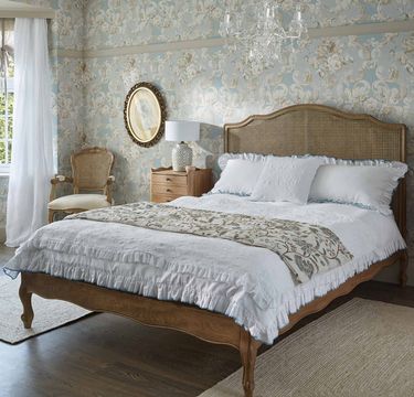 Elegant bedroom with a wooden bed, floral wallpaper, chandelier, and vintage chair. Soft lighting and a window with sheer curtains complete the look.