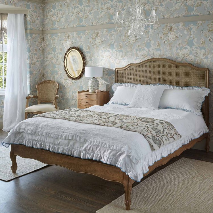 Elegant bedroom with a wooden bed, floral wallpaper, chandelier, and vintage chair. Soft lighting and a window with sheer curtains complete the look.
