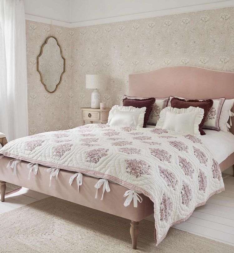 Elegant bedroom with a pink upholstered bed, floral quilt, decorative pillows, and a vintage mirror on a floral wallpapered wall. Soft natural light.