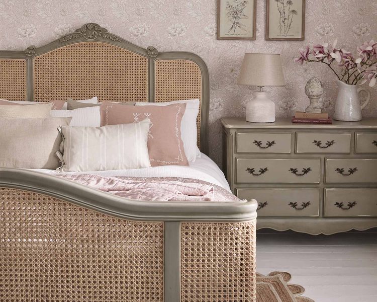 Vintage-style bedroom with a wicker bed, floral wallpaper, and a dresser with a lamp and floral decor. Soft pink and beige tones create a cozy ambiance.