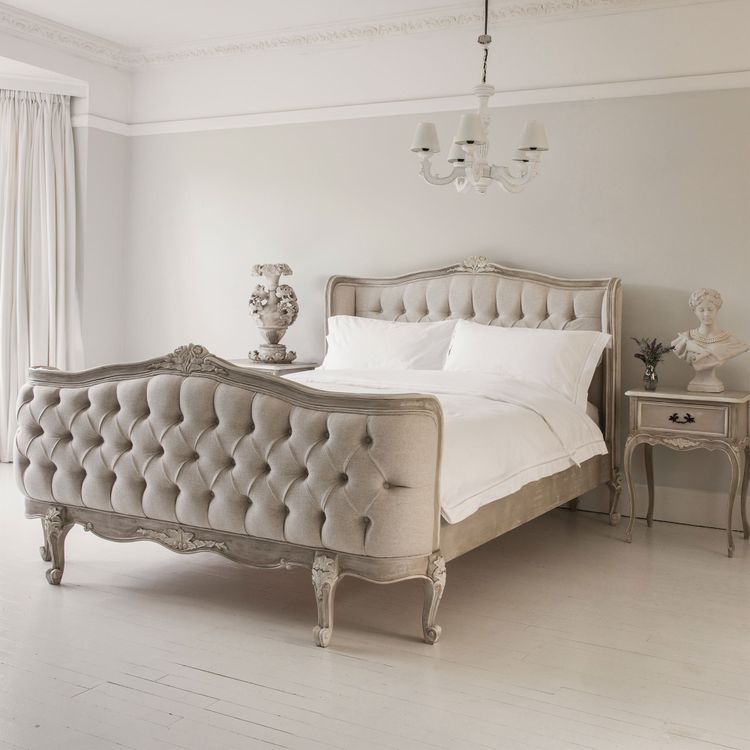 Elegant bedroom with a tufted, vintage-style bed, white bedding, a classic nightstand, a decorative vase, and a chandelier.
