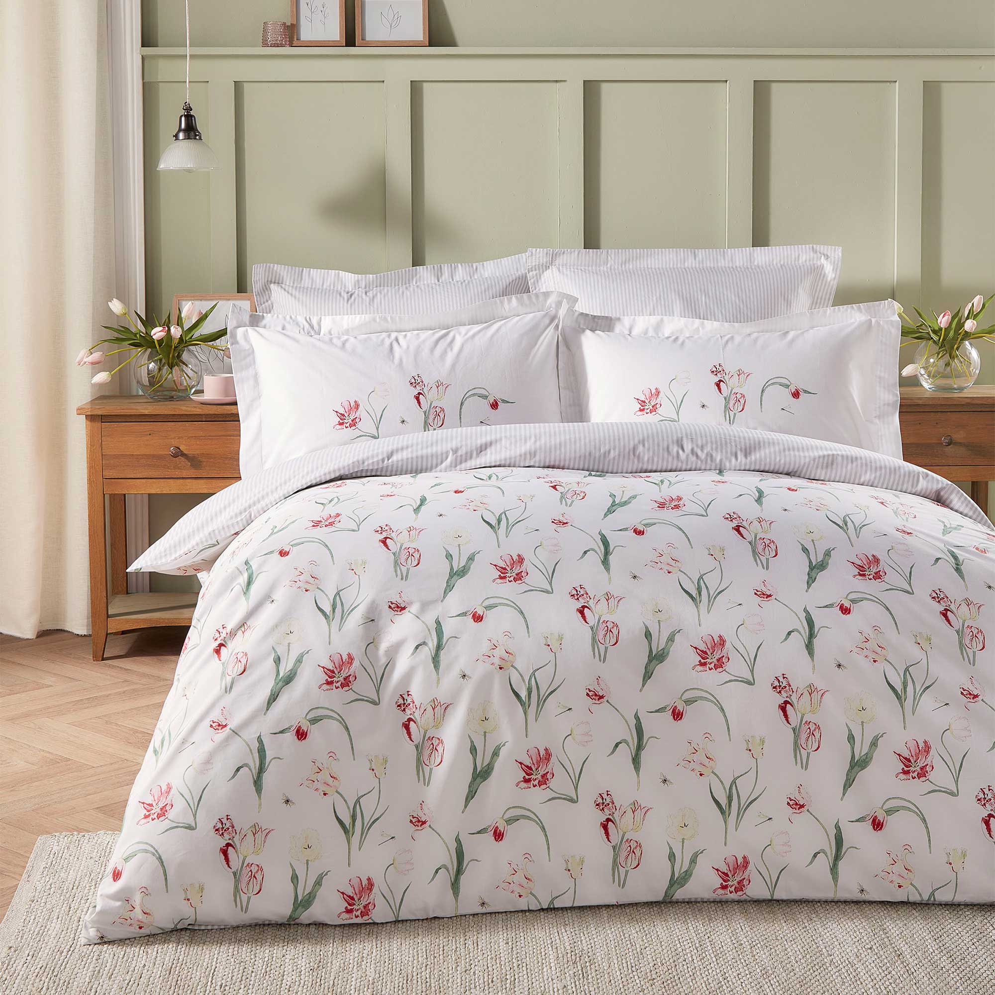 A neatly made bed with floral-patterned bedding, white pillows, and a wooden headboard. A wooden nightstand holds a lamp and flowers.