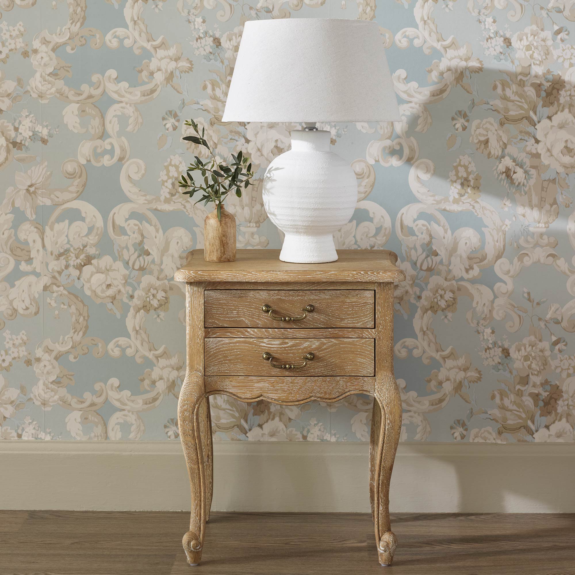 Vintage wooden side table with two drawers, a white lamp, and a small vase with greenery, set against floral wallpaper.